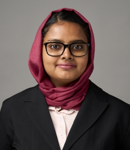 Woman with glasses, wearing red hijab, white shirt, black jacket, smiling. 