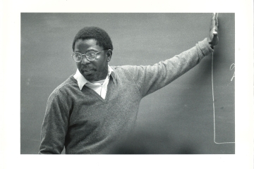 Man in glasses wearing sweater gesturing at chalkboard