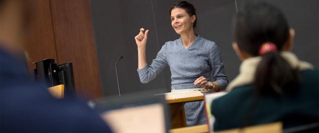 Professor Maeve Glass gestures in front of a blackboard in front of a lecture hall.