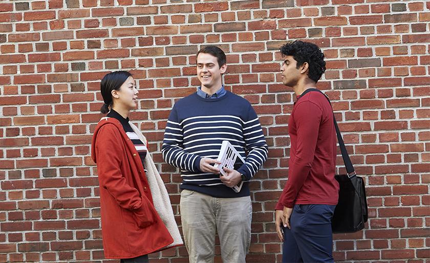 Three students stand and talk in front of a red brick wall.