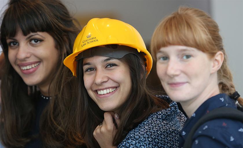 Three students smile, one of whom is wearing a yellow hardhat emblazoned with the Columbia Law logo.
