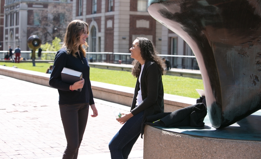 Two students talking near a statue on Revson Plaza.