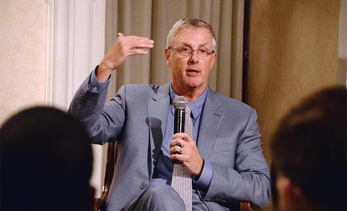 Bruce Sewell holds a microphone and gestures with his right hand.