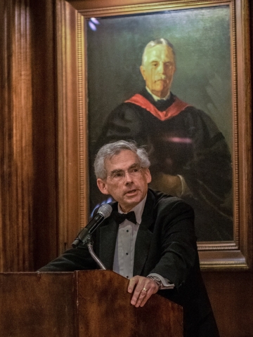 Man in bowtie at podium in front of painting of old white man