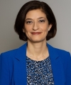 Pictured above is Caline Mouawad. She has dark brown bob. Silver studs and a silver neckless hang off her ears and neck. She is wearing a royal blue blazer and a patterned blouse. She has brown eyes. 