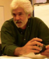 Pictured above is Kim Hopper. He is wearing an olive colored button up. He has short white facial hair and medium length white hair. 