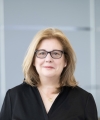 A white woman with blonde hair wears a black shirt and black-framed glasses.
