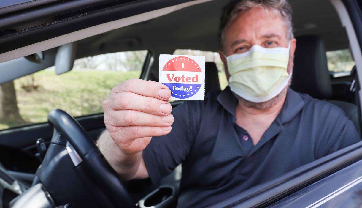 Man wearing a surgical mask for COVID-19 pandemic holds a sticker that says "I VOTED"