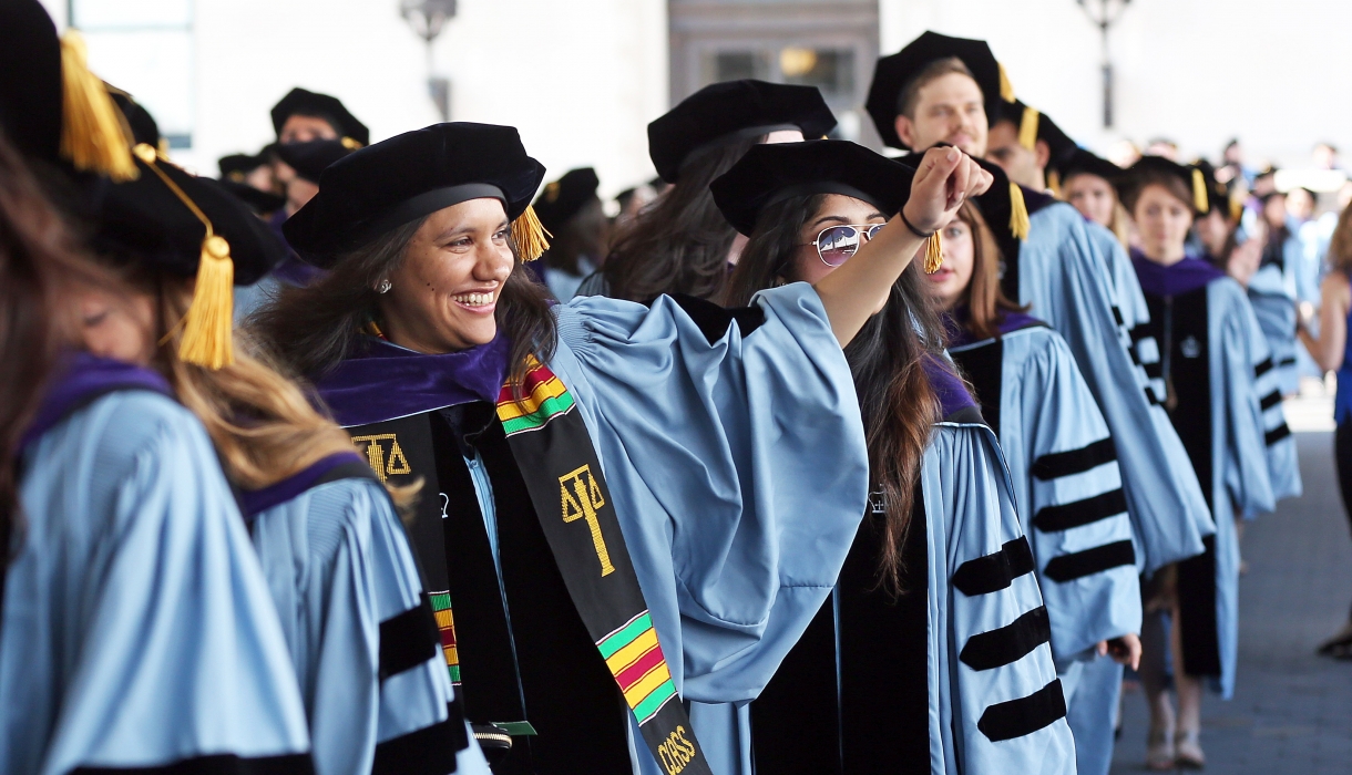 A student wearing academic regalia waves from the processional at the 2017 Graduation.