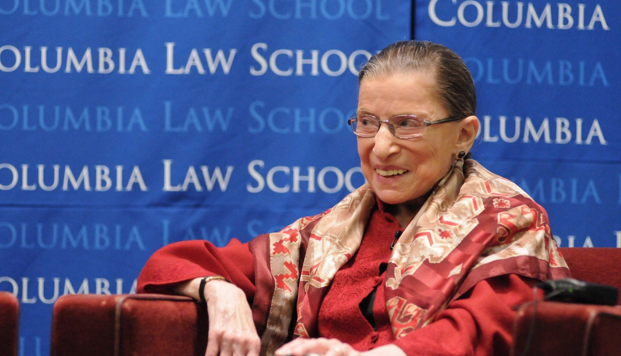 Ruth Bader Ginsburg smiling while wearing a red suit in front of a blue Columbia backdrop.