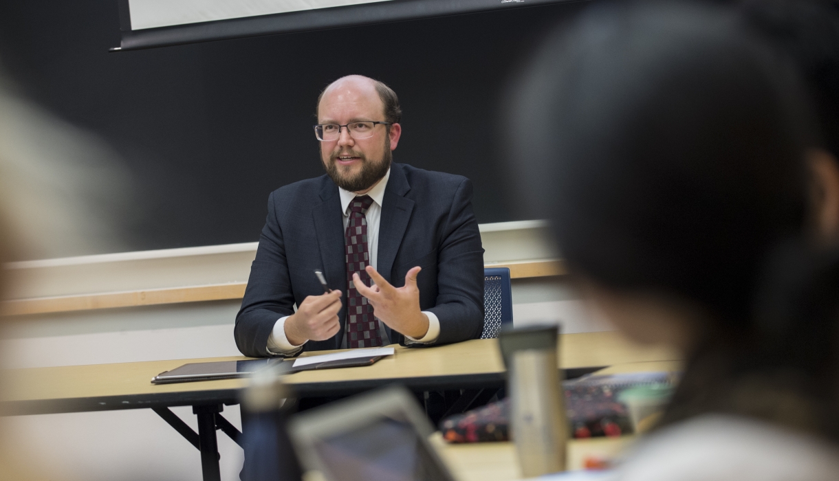 Man in glasses and suit at table in front of a classroom