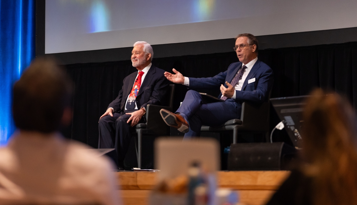 Two men seated onstage for panel discussion, both in suits. 