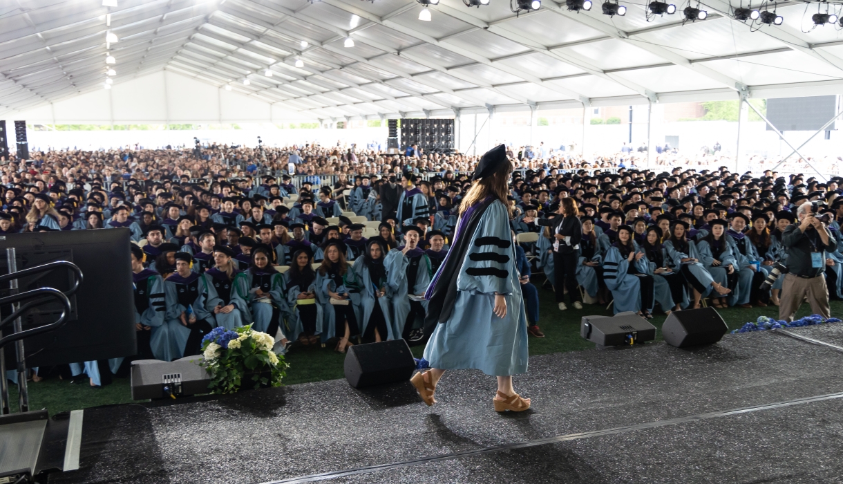 Woman in academic regalia crosses stage before large audience under enormous tent