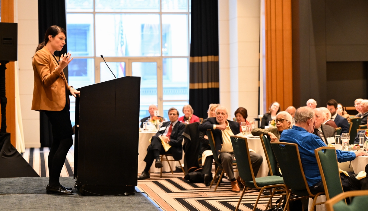 Woman standing at podium speaking to audience seated at tables