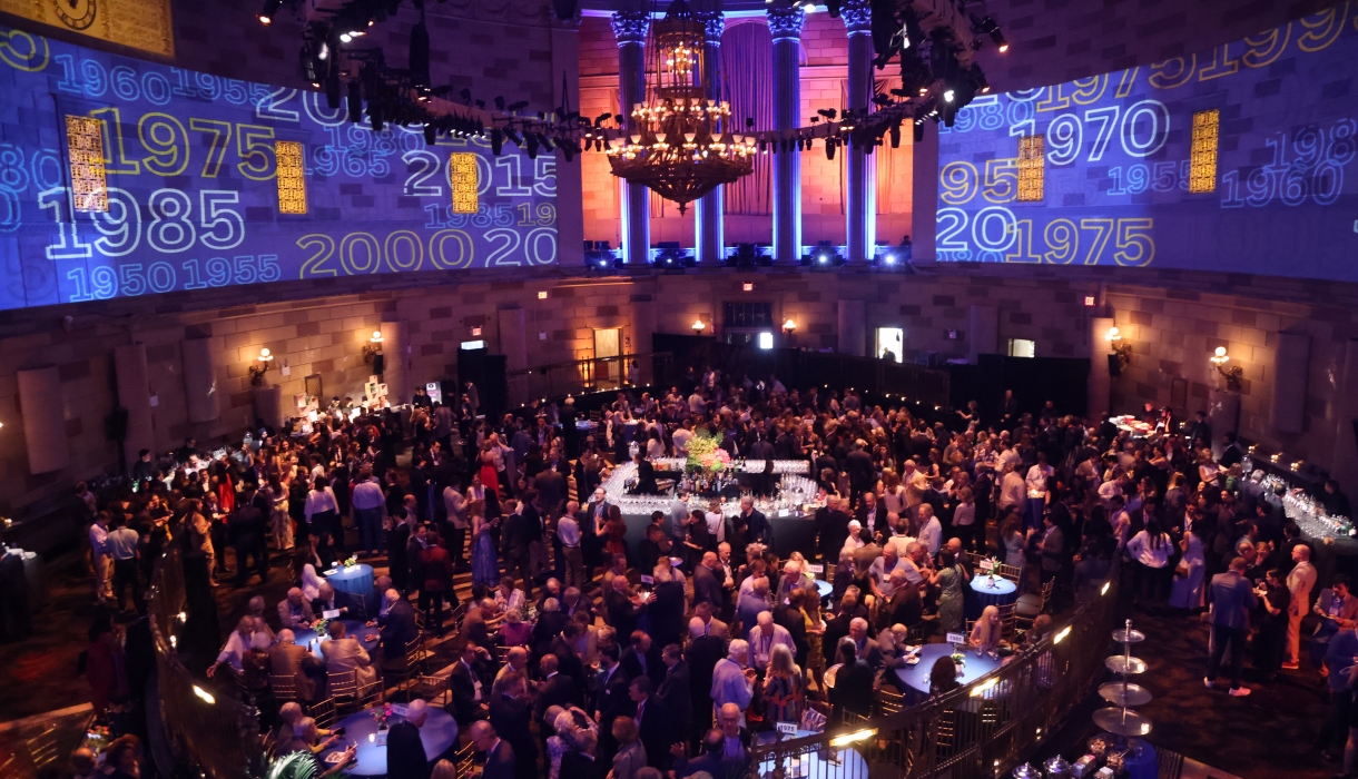 Wide shot of room filled with people at a cocktail party with Columbia logo projections on the walls. 