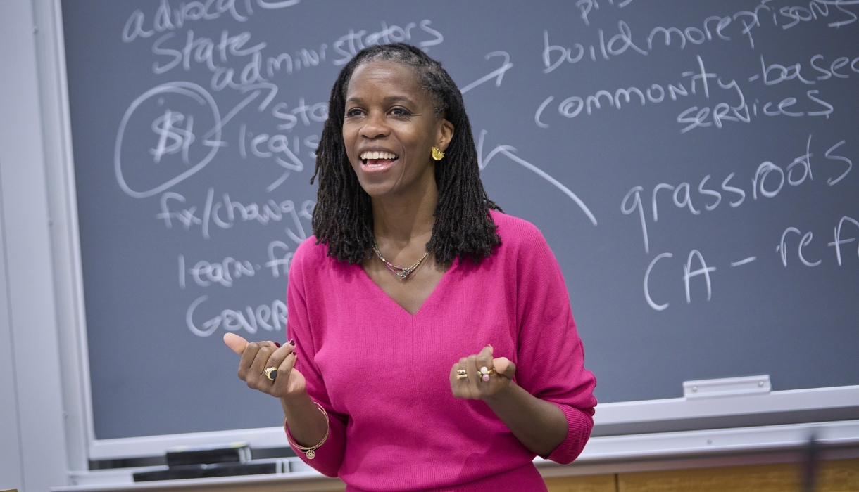 Olatunde Johnson in bright pink dress standing before blackboard covered in writing