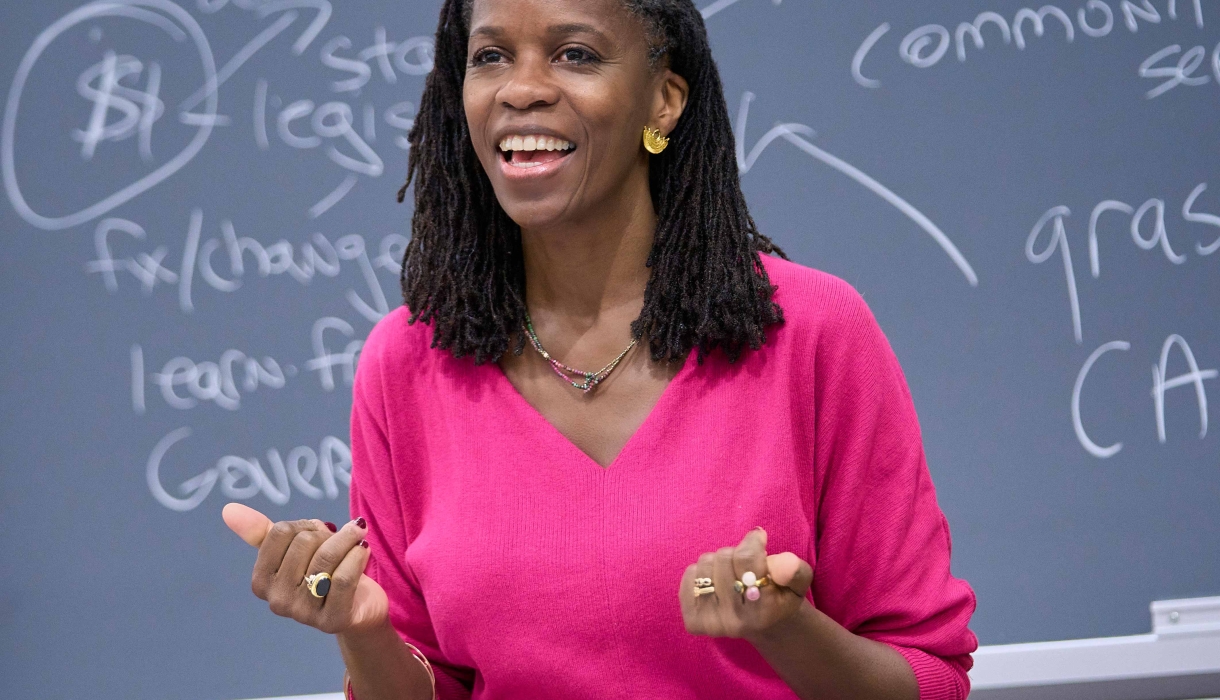 Olatunde Johnson in bright pink dress standing before blackboard covered in writing