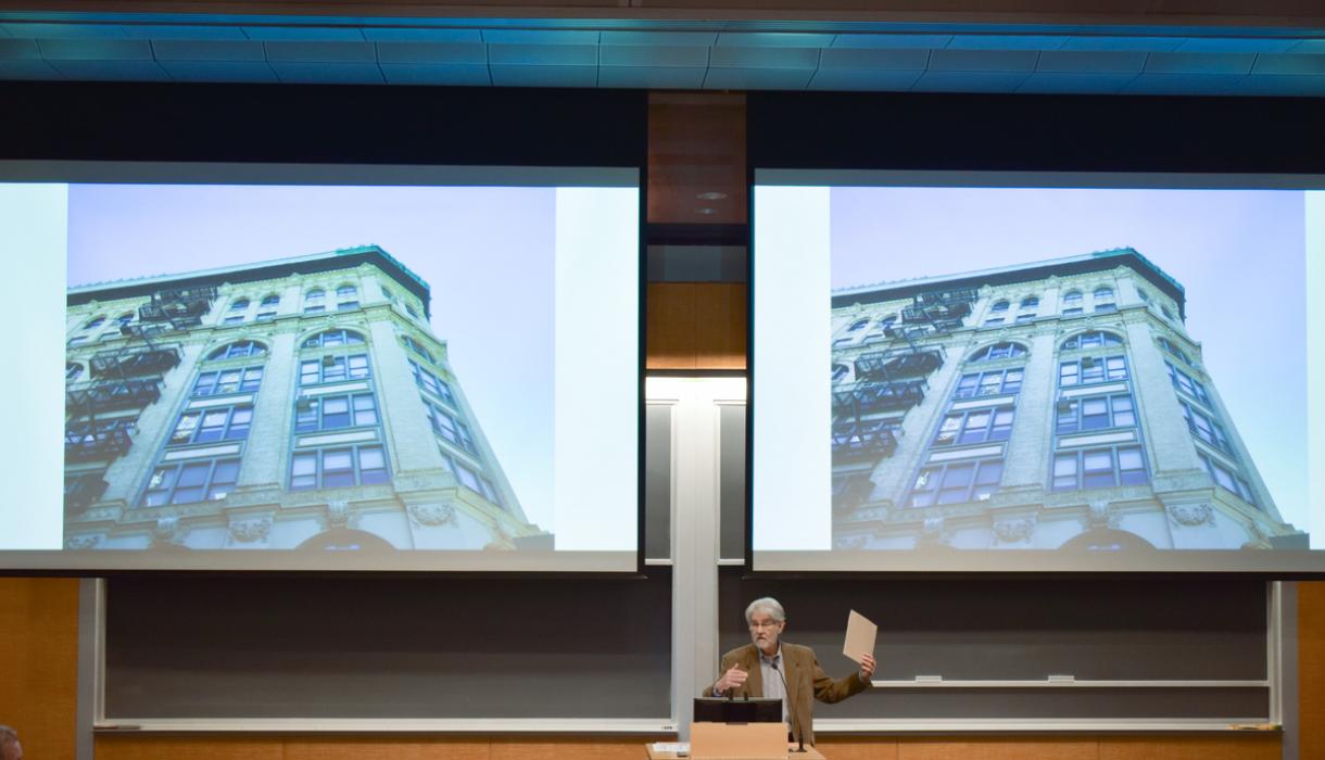 Professor Vincent Blasi gestures in front of a screen featuring a multistory building.