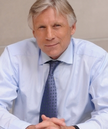 President Lee Bollinger smiles while folding his hands on a desk in front of him.