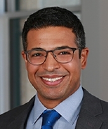 Smiling dark haired man with glasses in gray suit with blue tie
