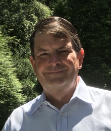 Thomas W. White picture above, stands smiling in front of grove of pine trees. He is wearing a baby blue button up. He has slightly combed over, short brown hair and fair skin. 