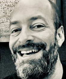 Pictured above is a black and white image of Andrew Friedman. He is seen donning a wide smile and black crew neck t-shirt. 