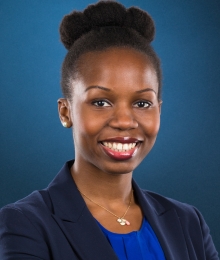 Pictured above is Temitope K. Yusuf. She is wearing a royal blue shirt under a navy blue blazer. She has pearl studs in each ear. She is smiling wide, and has wine colored rouge on her lips. She has dark brown eyes and curly hair tied back into a bun. 