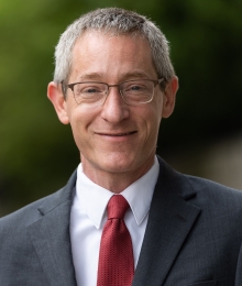 Professor Josh Gupta-Kagan, wearing glasses, a grey jacket, and a red necktie, smiles.