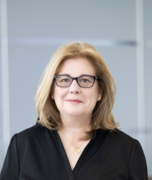 A white woman with blonde hair wears a black shirt and black-framed glasses.