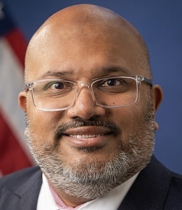 Man with gray beard wearing clear glasses before an American flag