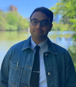 Man wearing glasses, a denim jacket, white shirt, and dark tie