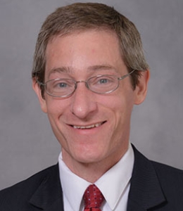 Headshot of a man wearing glasses, a white shirt, black blazer, and red tie