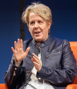 Woman speaking and gesturing while seated in a chair onstage. 