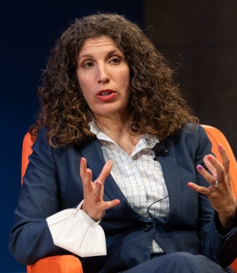Woman speaking and gesturing while seated in a chair onstage
