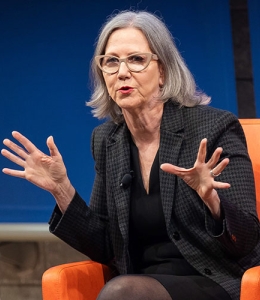 Woman speaking and gesturing seated in a chair onstage 