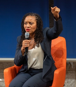 Woman speaking and gesturing while seated in a chair onstage