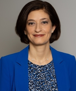 Pictured above is Caline Mouawad. She has dark brown bob. Silver studs and a silver neckless hang off her ears and neck. She is wearing a royal blue blazer and a patterned blouse. She has brown eyes.