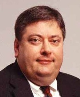 Pictured above is Kenneth C. Kettering. He has dark brown hair and eyes. He is wearing a black suit with a decorative tie.