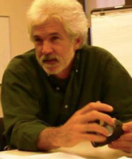 Pictured above is Kim Hopper. He is wearing an olive colored button up. He has short white facial hair and medium length white hair.