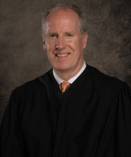 Pictured above is Richard J. Sullivan in traditional judge attire. He is wearing a white button up and copper tie. He has fair skin and grey hair. He is smiling wide. 