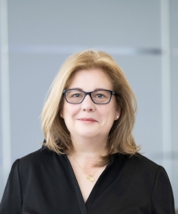 A white woman with blonde hair wears a black shirt and black-framed glasses.