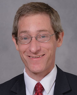 Headshot of a man wearing glasses, a white shirt, black blazer, and red tie