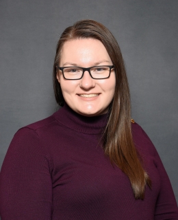 white woman with brown straight hair, purple sweater, and dark glasses smiles