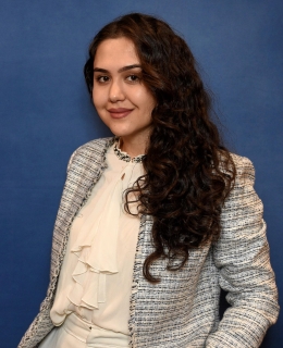 woman in a tweed blazer, whit blouse, and white pants with long brown curly hair smiles