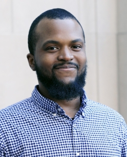 man with a beard and short dark hair in a blue checkered collared shirt smiles