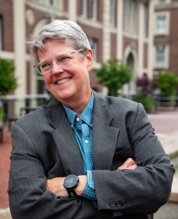 Image of Katherine Franke, looking away while smiling, with arms crossed, wearing a grey suit and blue shirt