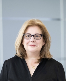 A white woman with blonde hair wears a black shirt and black-framed glasses.
