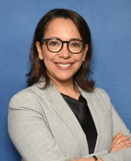 Woman with shoulder-length brown hair with a slight curl at the bottom, large glasses, in a gray blazer and black top smiles