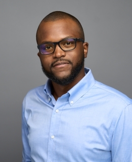 man with shaved head in glasses and a light blue collared shirt smiles