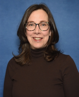 woman with medium loose wavey hair, round glasses, and brown turtleneck top smiles; there is a blue background